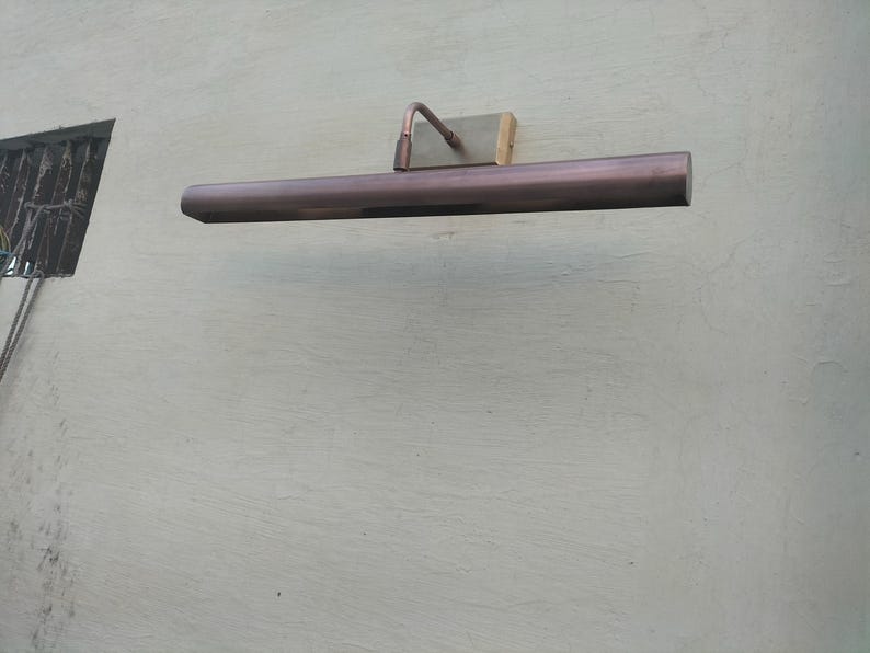May include: A horizontal, tubular picture light with a metallic copper finish. The fixture is mounted to a wall via a rectangular, brushed-metal base plate connected by a curved, adjustable arm that extends from the center of the light tube.