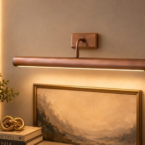 May include: A wall-mounted picture light with a cylindrical, brushed copper-toned metal finish. The fixture features a rectangular base plate and a horizontal arm that extends to a long, tubular lamp head, casting a warm, downward-facing glow.