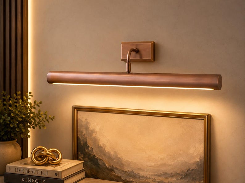 May include: A wall-mounted picture light with a cylindrical, brushed copper-toned metal finish. The fixture features a rectangular base plate and a horizontal arm that extends to a long, tubular lamp head, casting a warm, downward-facing glow.