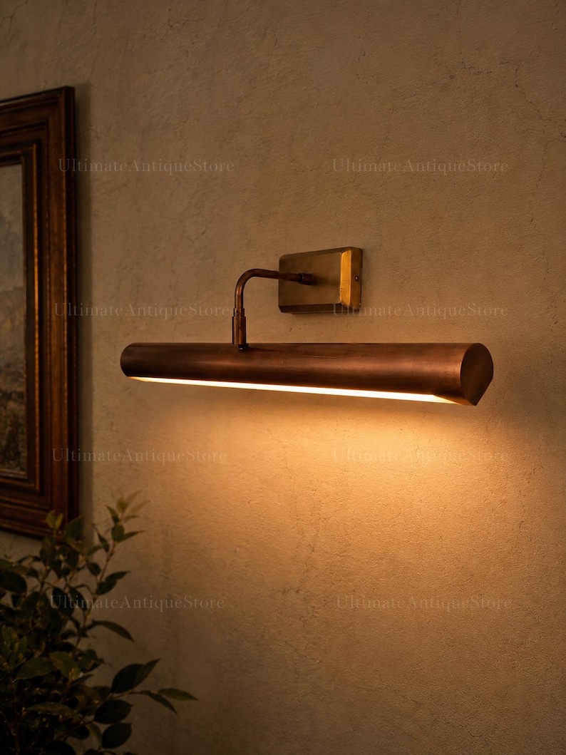 May include: A wall-mounted picture light with a cylindrical, metallic copper-toned shade and a rectangular brass-coloured base. The fixture features a curved arm connecting the shade to the wall plate, emitting a warm, downward-facing light.