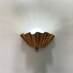 Könnte beinhalten: Eine goldfarbene Wandleuchte mit einem muschelartigen Design. Die Leuchte hat einen abgerundeten Sockel und strahlt helles Licht aus. Die Leuchte ist an einer weißen Wand befestigt.