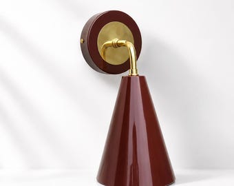 May include: A wall-mounted lamp featuring a deep burgundy, conical shade and a circular wall plate in the same color. The fixture includes a polished gold-tone metal arm that connects the shade to the wall plate, creating a sleek, modern look.