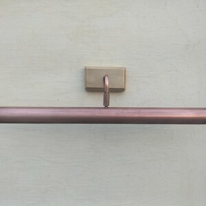 May include: A copper-toned metal picture light featuring a long, cylindrical horizontal shade attached to a rectangular wall-mounting plate by a single curved arm. The fixture has a smooth, metallic finish and a clean, linear design.