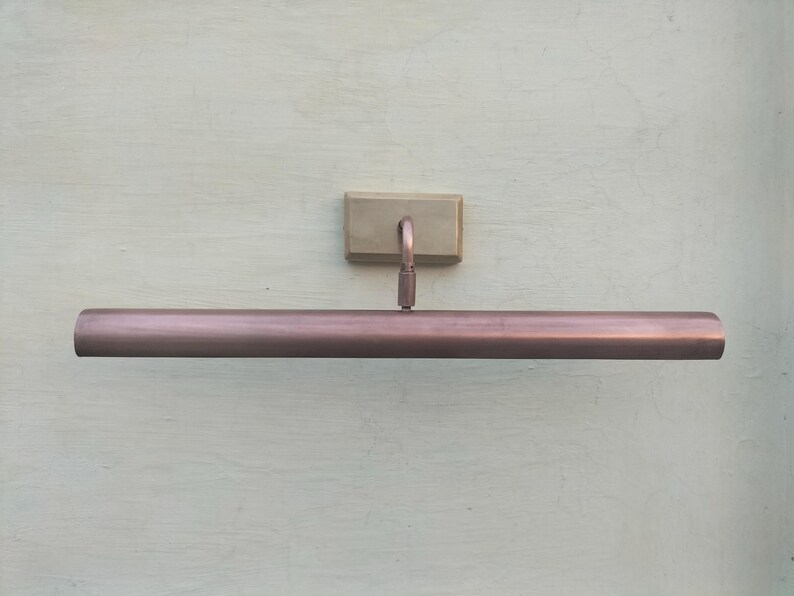 May include: A copper-toned metal picture light featuring a long, cylindrical horizontal shade attached to a rectangular wall-mounting plate by a single curved arm. The fixture has a smooth, metallic finish and a clean, linear design.