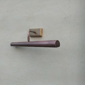 May include: A wall-mounted picture light with a cylindrical, dark bronze-toned metal shade. The fixture features a rectangular mounting plate and a curved arm connecting the shade to the base, providing a sleek, functional design for illumination.