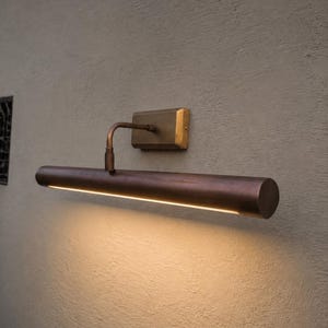 May include: A wall-mounted picture light with a cylindrical, dark bronze-toned metal housing and a rectangular brass-colored base plate. The fixture features a curved arm connecting the base to the light tube, which emits a warm downward glow.