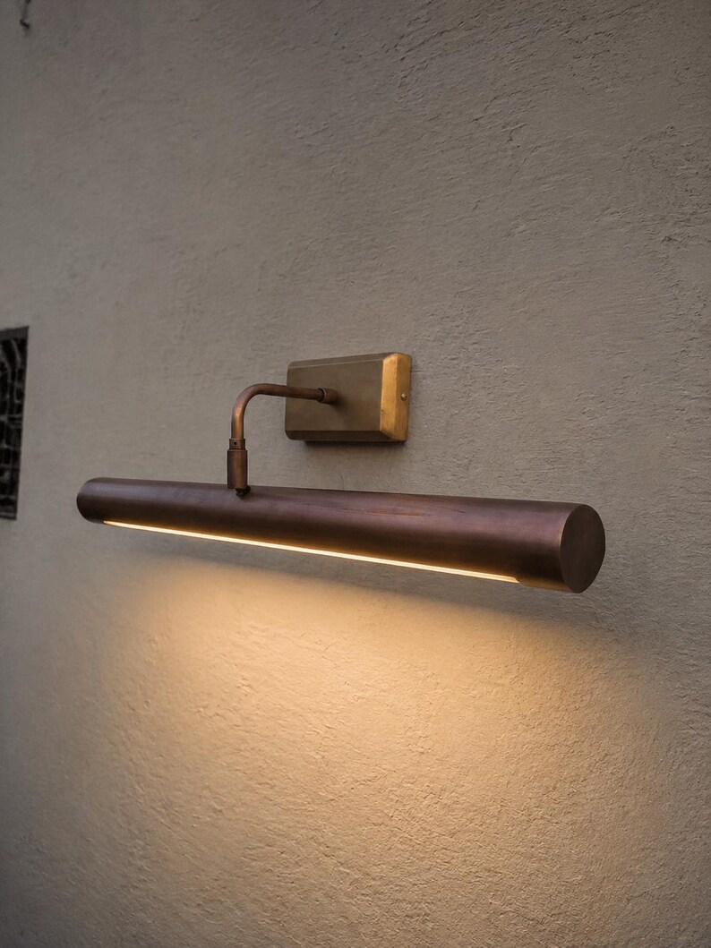 May include: A wall-mounted picture light with a cylindrical, dark bronze-toned metal housing and a rectangular brass-colored base plate. The fixture features a curved arm connecting the base to the light tube, which emits a warm downward glow.