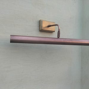 May include: A wall-mounted picture light fixture featuring a long, cylindrical, metallic copper-toned shade attached to a rectangular brass-colored base plate by a single adjustable arm. The fixture has a smooth, polished finish throughout.
