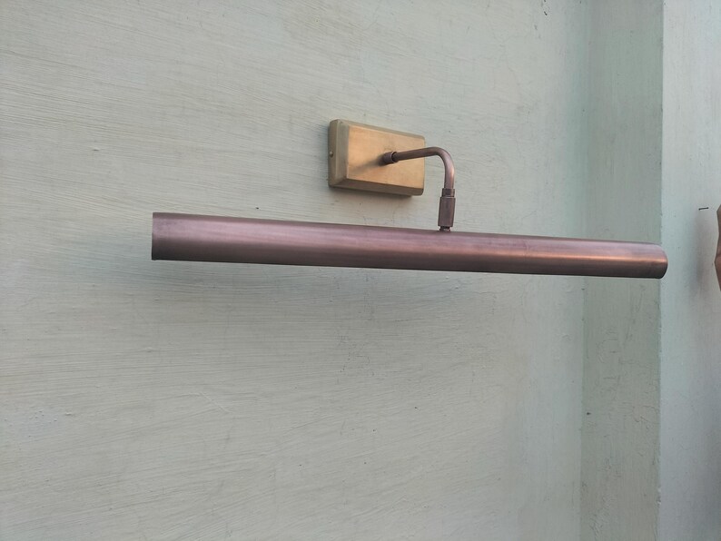 May include: A wall-mounted picture light fixture featuring a long, cylindrical, metallic copper-toned shade attached to a rectangular brass-colored base plate by a single adjustable arm. The fixture has a smooth, polished finish throughout.