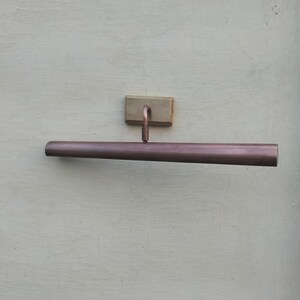 May include: A wall-mounted picture light with a long, cylindrical, metallic copper-toned shade. The fixture features a rectangular brass-colored mounting plate and a short, adjustable arm connecting the shade to the base for directional lighting.