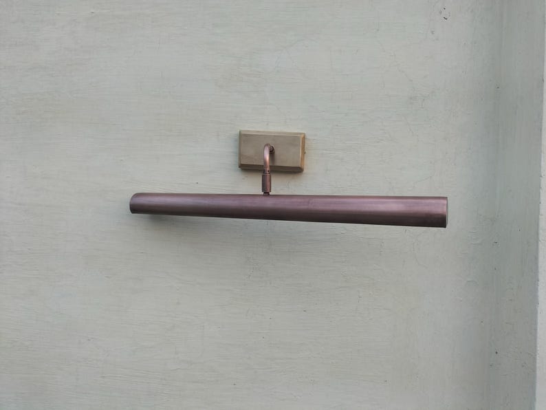 May include: A wall-mounted picture light with a long, cylindrical, metallic copper-toned shade. The fixture features a rectangular brass-colored mounting plate and a short, adjustable arm connecting the shade to the base for directional lighting.