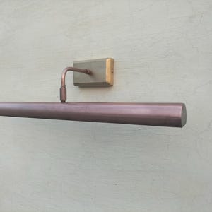 May include: A wall-mounted picture light with a long, cylindrical, metallic copper-toned shade. The fixture features a rectangular base plate and a curved arm that connects the base to the center of the horizontal light tube, extending outward.