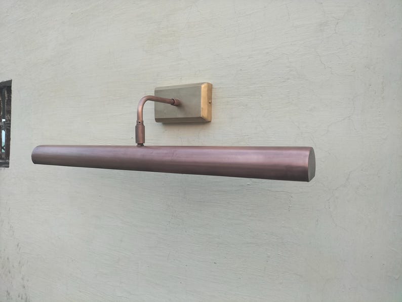 May include: A wall-mounted picture light with a long, cylindrical, metallic copper-toned shade. The fixture features a rectangular base plate and a curved arm that connects the base to the center of the horizontal light tube, extending outward.
