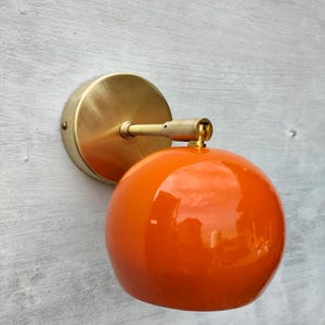 Könnte beinhalten: Eine Wandleuchte mit einem glänzenden, kugelförmigen orangefarbenen Glasschirm, der an einer kreisförmigen Basis aus gebürstetem Messing befestigt ist. Die Lampe verfügt über einen kurzen, horizontalen Messingarm, der die Basis mit der orangefarbenen Kugel verbindet und eine feste Lichtquelle mit polierter Metalloberfläche und einem leuchtend einfarbigen Glaselement bietet.