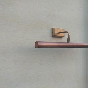 May include: A wall-mounted picture light with a long, cylindrical copper-toned shade and a rectangular brass-colored base. The fixture features a thin, curved arm connecting the base to the center of the horizontal light housing.