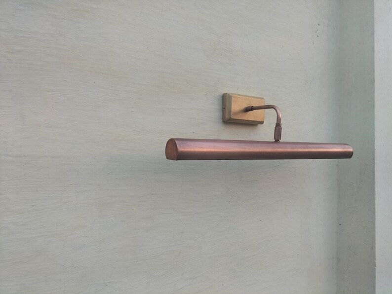 May include: A wall-mounted picture light with a long, cylindrical copper-toned shade and a rectangular brass-colored base. The fixture features a thin, curved arm connecting the base to the center of the horizontal light housing.