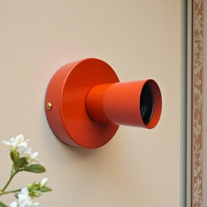 May include: A vibrant orange wall sconce with a cylindrical light fixture. The sconce has a circular base and a tubular arm extending outward. The fixture is mounted on a beige wall, with a small brass screw visible. The light opening is black.