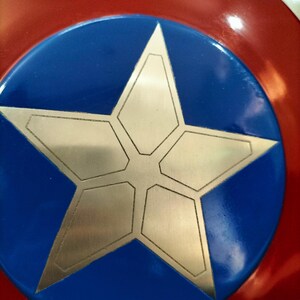 Op de afbeelding: Een rood, wit en blauw schild met een grote zilveren ster in het midden. Het schild heeft een cirkelvormig ontwerp met een blauw midden en een rode buitenring. De ster is gemaakt van geometrische vormen.