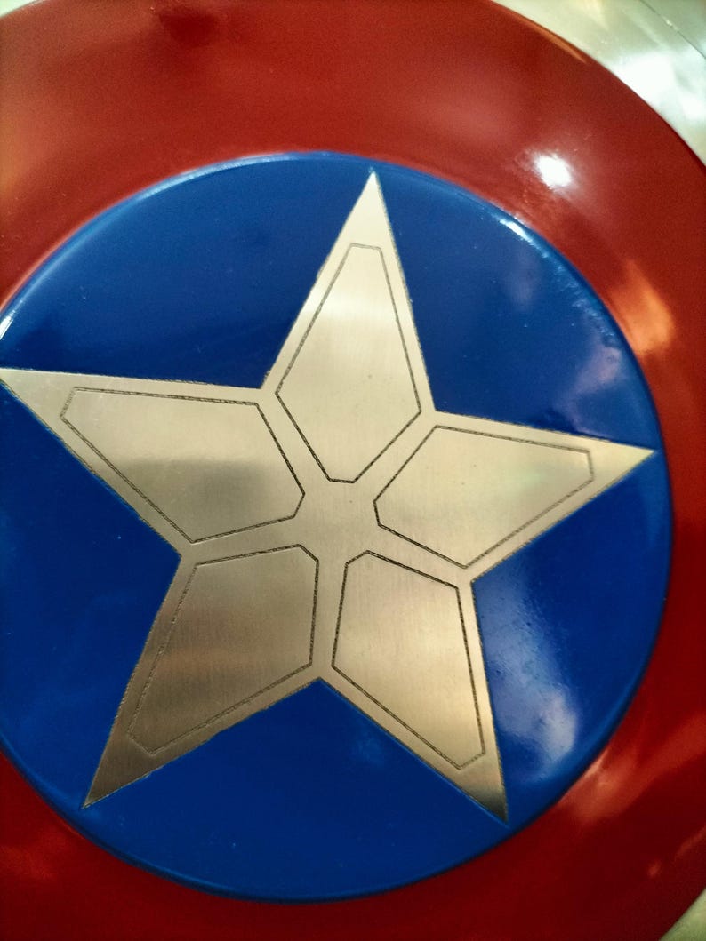 Op de afbeelding: Een rood, wit en blauw schild met een grote zilveren ster in het midden. Het schild heeft een cirkelvormig ontwerp met een blauw midden en een rode buitenring. De ster is gemaakt van geometrische vormen.