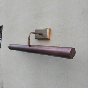 May include: A wall-mounted picture light with a long, cylindrical, dark bronze-toned metal shade. The fixture features a rectangular mounting plate in a brushed brass finish, connected to the shade by a single, adjustable curved metal arm.