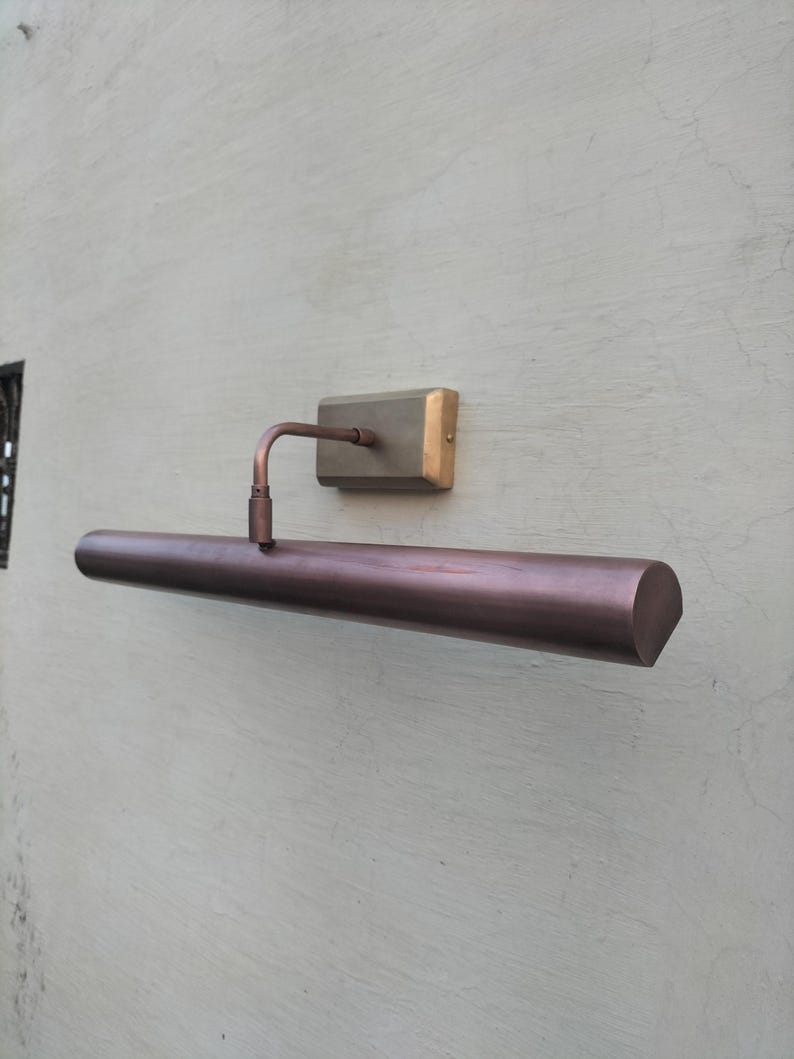 May include: A wall-mounted picture light with a long, cylindrical, dark bronze-toned metal shade. The fixture features a rectangular mounting plate in a brushed brass finish, connected to the shade by a single, adjustable curved metal arm.