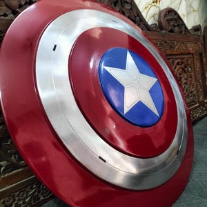 Op de afbeelding: Een rood, wit en blauw schild met een starembleem. Het schild heeft een glanzende afwerking en een zilverkleurige ring rond het midden. Het schild is een replica van Captain America's schild.