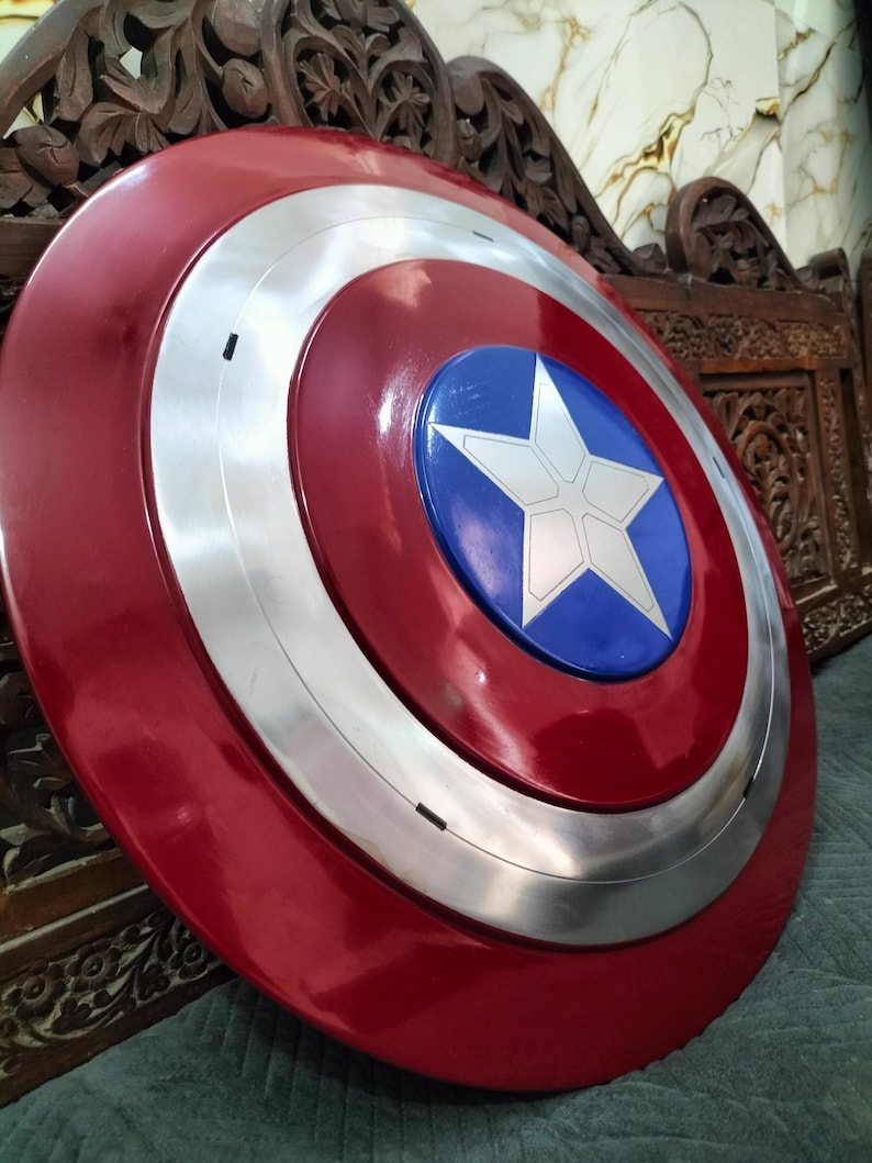Op de afbeelding: Een rood, wit en blauw schild met een starembleem. Het schild heeft een glanzende afwerking en een zilverkleurige ring rond het midden. Het schild is een replica van Captain America's schild.