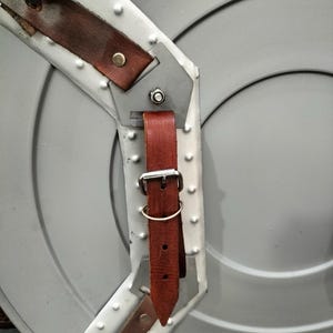 Op de afbeelding: Een grijs metalen schild met een geklonken rand en bruine leren riemen. Het schild heeft een cirkelvormig ontwerp met concentrische ringen. De leren riemen zijn vastgezet met metalen gespen en klinknagels.