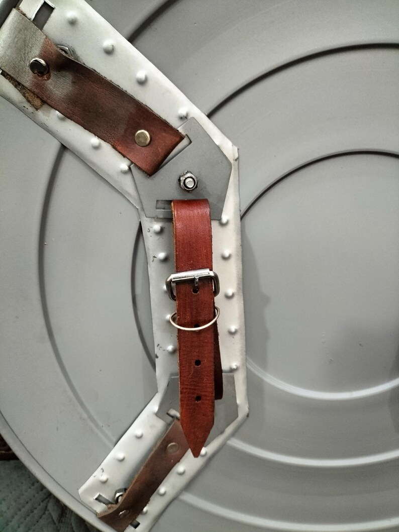 Op de afbeelding: Een grijs metalen schild met een geklonken rand en bruine leren riemen. Het schild heeft een cirkelvormig ontwerp met concentrische ringen. De leren riemen zijn vastgezet met metalen gespen en klinknagels.