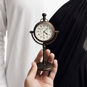 May include: A vintage-style brass clock with a white face and black numerals. The clock is held in front of a white shirt and black fabric background. The clock's hands indicate a time of approximately 2:50.