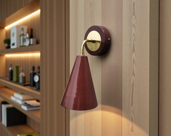 May include: A wall-mounted sconce featuring a conical, deep red shade and a polished brass arm. The fixture is attached to a circular, dark red base plate mounted on a vertical wood-paneled wall, casting a warm light downward.