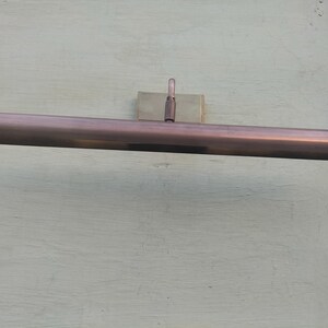 May include: A horizontal, rectangular picture light fixture with a metallic copper finish. The elongated, tubular lamp head is attached to a central mounting bracket by a single, adjustable vertical arm, designed for wall-mounted illumination.