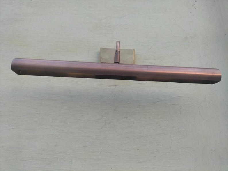 May include: A horizontal, rectangular picture light fixture with a metallic copper finish. The elongated, tubular lamp head is attached to a central mounting bracket by a single, adjustable vertical arm, designed for wall-mounted illumination.
