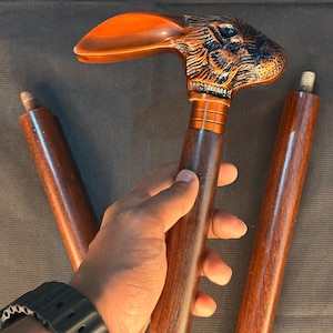 May include: A wooden walking cane with a carved rabbit head handle. The handle is a mix of brown and orange tones, with detailed carving. The cane shaft is a dark brown color, and there are two additional cane sections.