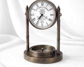 May include: A bronze-toned metal desk clock featuring a white face with black Roman numerals, the text "VICTORIA LONDON" and "1876", and a ship's wheel and anchor graphic. The clock is suspended between two pillars above a circular compass base.