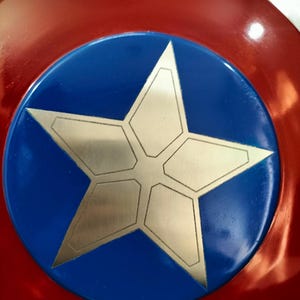 Op de afbeelding: Een rond schild met een rode buitenrand, een blauwe centrale cirkel en een zilveren sterontwerp. Het schild heeft een glanzende afwerking en is waarschijnlijk een verzamelobject of cosplay-item.