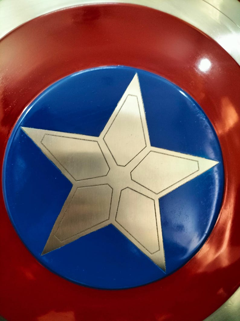 Op de afbeelding: Een rond schild met een rode buitenrand, een blauwe centrale cirkel en een zilveren sterontwerp. Het schild heeft een glanzende afwerking en is waarschijnlijk een verzamelobject of cosplay-item.