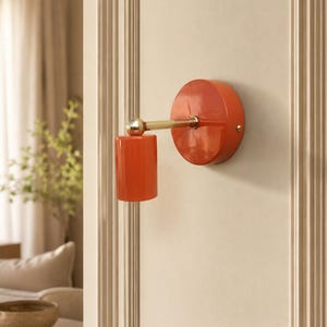 May include: A wall-mounted light fixture featuring a circular orange base and a matching cylindrical orange shade. The shade is connected to the base by a horizontal brass-colored metal arm with a spherical joint, allowing for adjustable positioning.