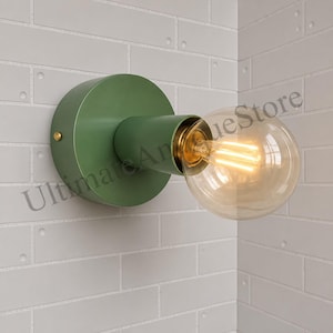 Może przedstawiać: Zielona lampa ścienna z okrągłą podstawą i cylindrycznym ramieniem. Posiada dużą, przezroczystą, okrągłą żarówkę z ciepłym, świecącym żarnikiem. Oprawa jest zamontowana na białej ceglanej ścianie. Widoczny jest tekst "Ultimate Antique Store".