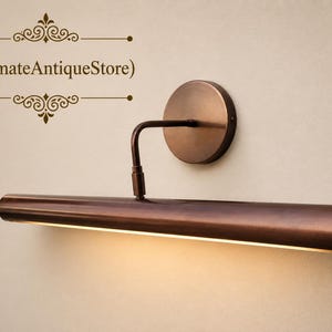 May include: A bronze-colored picture light fixture with a circular wall mount and a rectangular light bar. The light bar extends horizontally, emitting a warm glow. The fixture is mounted on a neutral-colored wall. The text "(UltimateAntiqueStore)" is visible.