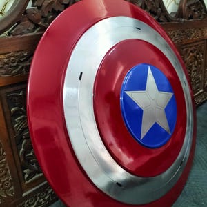 Op de afbeelding: Een rood, wit en blauw schild met een starembleem. Het schild heeft een glanzende afwerking en een cirkelvormig ontwerp. Het centrale embleem is een witte ster op een blauwe achtergrond, omringd door zilveren en rode concentrische cirkels.