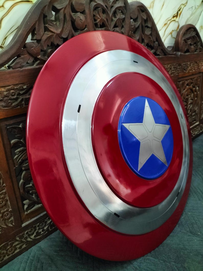 Op de afbeelding: Een rood, wit en blauw schild met een starembleem. Het schild heeft een glanzende afwerking en een cirkelvormig ontwerp. Het centrale embleem is een witte ster op een blauwe achtergrond, omringd door zilveren en rode concentrische cirkels.