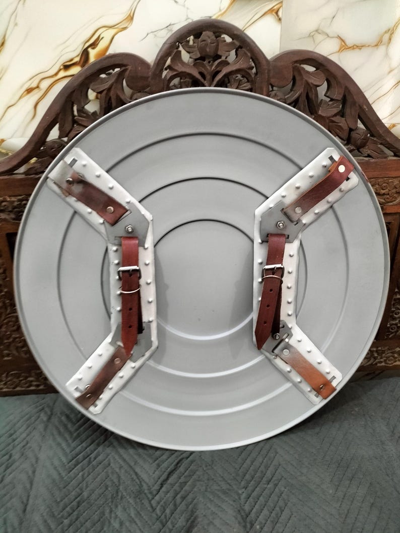 Op de afbeelding: Een grijze, cirkelvormige schild met concentrische ringen en geklonken metalen beugels. Bruine leren riemen met zilveren gespen zijn aan de beugels bevestigd. Het schild heeft een verweerde uitstraling.