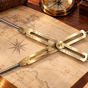 May include: A brass drafting compass with two adjustable arms featuring pointed metal tips. The tool has engraved markings and the text CIRCLES and LINES. It is positioned on a vintage-style map with a compass rose and grid lines.