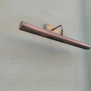 May include: A horizontal, tubular picture light with a metallic copper finish. The fixture features a rectangular wall-mounting plate and a thin, angled arm that connects to the center of the long, cylindrical light housing, extending outward.