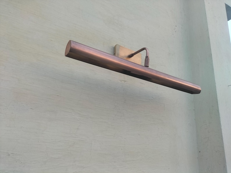 May include: A horizontal, tubular picture light with a metallic copper finish. The fixture features a rectangular wall-mounting plate and a thin, angled arm that connects to the center of the long, cylindrical light housing, extending outward.
