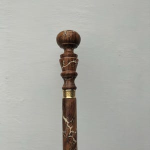 May include: A wooden walking cane with a dark brown finish and a decorative design. The cane features a rounded top, a detailed midsection, and a gold-colored band. The shaft has a unique, light-colored, lightning-like pattern.