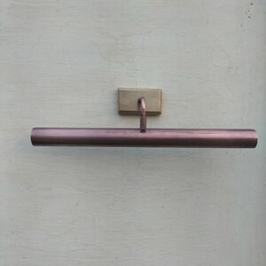 May include: A wall-mounted picture light featuring a long, cylindrical, metallic copper-toned shade. The fixture includes a rectangular, brushed brass-colored base plate and a short, curved arm that connects the base to the center of the tube.