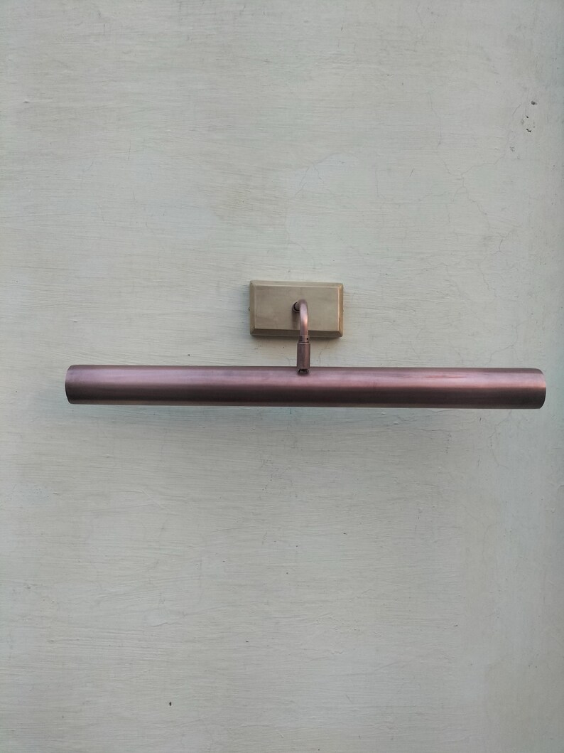 May include: A wall-mounted picture light featuring a long, cylindrical, metallic copper-toned shade. The fixture includes a rectangular, brushed brass-colored base plate and a short, curved arm that connects the base to the center of the tube.