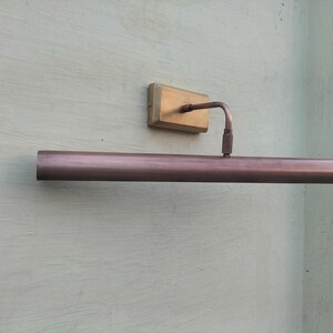 May include: A wall-mounted picture light with a long, cylindrical, metallic copper-toned shade. The fixture features a rectangular brass-colored base plate and a thin, curved arm that connects the base to the center of the horizontal light tube.